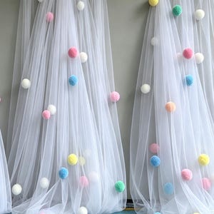 Puede incluir: Dos paneles de tela de tul blanco transparente drapeados verticalmente, con un surtido de pequeños pompones difusos en colores pastel, incluyendo rosa, azul, amarillo, verde y blanco, esparcidos por el material ligero y translúcido.