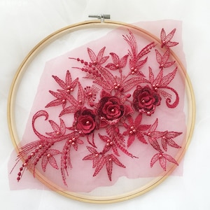 May include: Burgundy floral applique with three dimensional flowers and pearl accents. The applique is displayed on a wooden embroidery hoop.