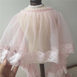 May include: A delicate pink tulle cape with a white lace trim. The cape is gathered at the neck and has a flowing, ethereal look.