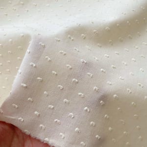 Puede incluir: Primer plano de una tela color crema con textura de puntos. La tela está doblada, mostrando la textura y la ligera transparencia del material. Los puntos están en relieve y espaciados uniformemente.