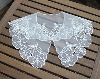 May include: White lace collar with a floral design. The collar is made of delicate fabric and has a scalloped edge.