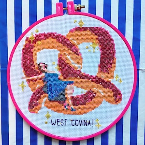 May include: A pink and white embroidery hoop with a cross-stitch design of a woman in a blue dress sitting on a pretzel. The design is surrounded by gold glitter stars and the text "West Covina!" is stitched in the bottom centre.
