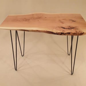 Peut inclure: Une table en bois avec un plateau à bord vif et des pieds en épingle à cheveux noirs. La table est en bois naturel de couleur brun clair et présente un motif de grain unique.