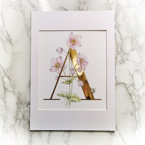 A es para Anemone Gilded Print. Letra A en pan de oro de 23,5 quilates con una anémona rosa entrelazada y un abejorro. Letra A con forma de flor.