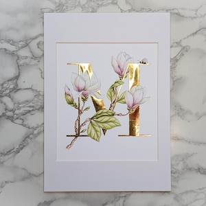 Estampado dorado con la letra M de Magnolia. Una letra M dorada con una magnolia entrelazada y un abejorro. Ideal para cualquier persona cuyo nombre comience con M.
