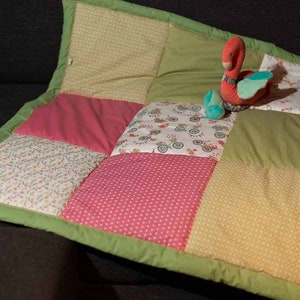 Könnte beinhalten: Eine Patchwork-Babydecke mit grünem Rand und einem Farbschema in Rosa, Weiß und Grün. Ein Plüsch-Schwan-Spielzeug in Rosa und Grün liegt auf der Decke.