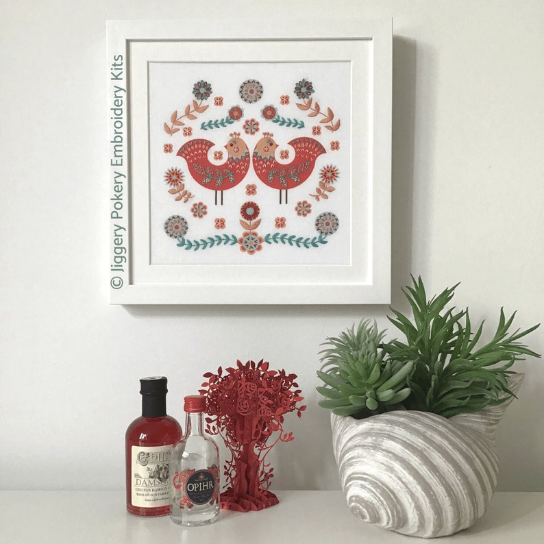 Folk art bird embroidery mounted and framed, shown hanging on a cream wall with miniature gin bottles for scale.