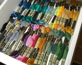 May include: A collection of embroidery floss skeins arranged in a drawer, featuring a wide spectrum of colors including blues, greens, yellows, oranges, purples, and pinks. Each skein is secured with a paper label displaying a barcode and brand logo.