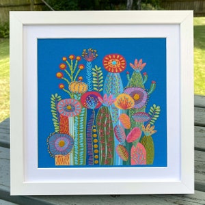 Cactus embroidery kit on blue fabric in a square white frame. Photo taken outdoors on a garden table