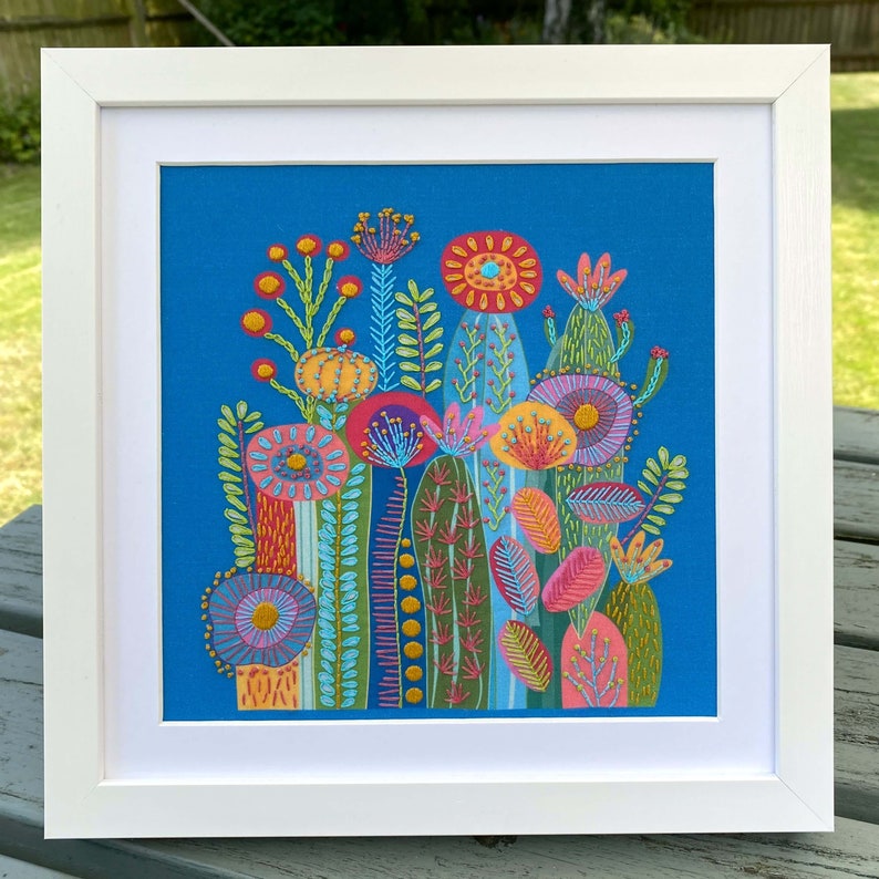 Cactus embroidery kit on blue fabric in a square white frame. Photo taken outdoors on a garden table