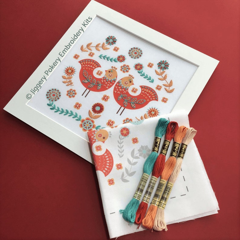 Completed folk art bird embroidery kit in white mount, shown with fabric with the embroidery design printed onto it and DMC threads in cream, light orange, red and turquoise.