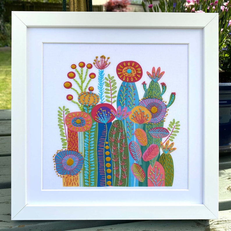 Cactus embroidery kit on white fabric in a square white frame. Photo taken outdoors on a garden table.