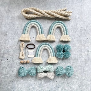Könnte beinhalten: Ein Bastelset zum Herstellen von drei Makramee-Regenbögen. Das Set enthält weißes Baumwollseil, blaues und weißes Garn, Holzperlen und Pompons.