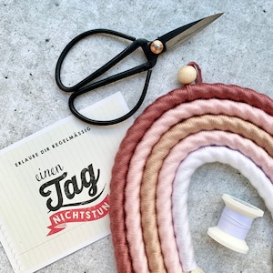 Op de afbeelding: Een handgemaakte macram&eacute; regenboog wandhanger in tinten van roze, beige en wit. De regenboog is gemaakt van garen en heeft een houten kraal aan de bovenkant. De afbeelding bevat ook een kaart met de tekst "Erlaube dir regelm&auml;ssig einen Tag Nichtstun" en een schaar.