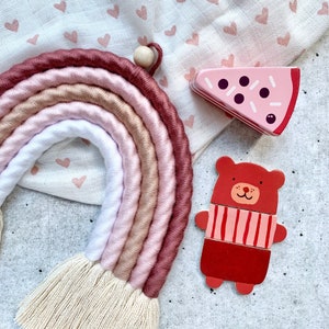 Op de afbeelding: Een roze en bruine regenboog garen wanddecoratie met een houten kraal aan de bovenkant. De regenboog hangt aan een witte stof met een hartjespatroon. Er is een houten speelgoedbeer en een houten speelgoedpizzapunt op de stof.