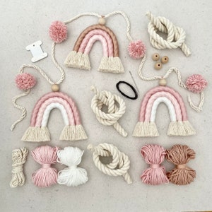 Könnte beinhalten: Ein Set an DIY Regenbogen-Wandbehängen mit rosa, weißem und beigefarbenem Garn. Das Set enthält Garn, Holzperlen und Schnur.