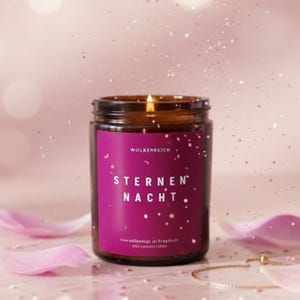 Könnte beinhalten: Eine brennende Kerze in einem braunen Glas mit einem lila Etikett. Auf dem Etikett steht "STERNEN NACHT" und "WOLKENREICH". Die Kerze ist von rosa Blütenblättern und Glitzer umgeben.