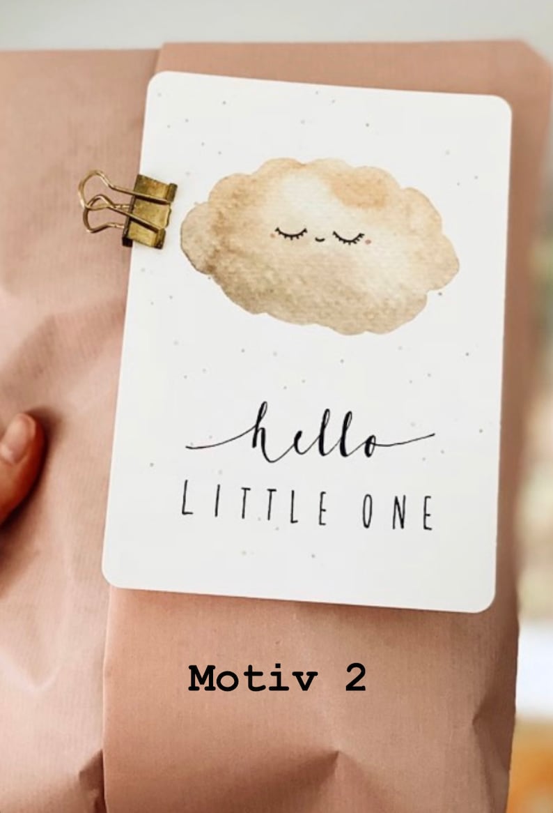 Op de afbeelding: Een witte kaart met een bruine aquarel wolk met een glimlachend gezicht en gesloten ogen. De kaart zegt "hello little one" in zwarte schrift. De kaart is bevestigd aan een bruine papieren zak met een gouden paperclip.