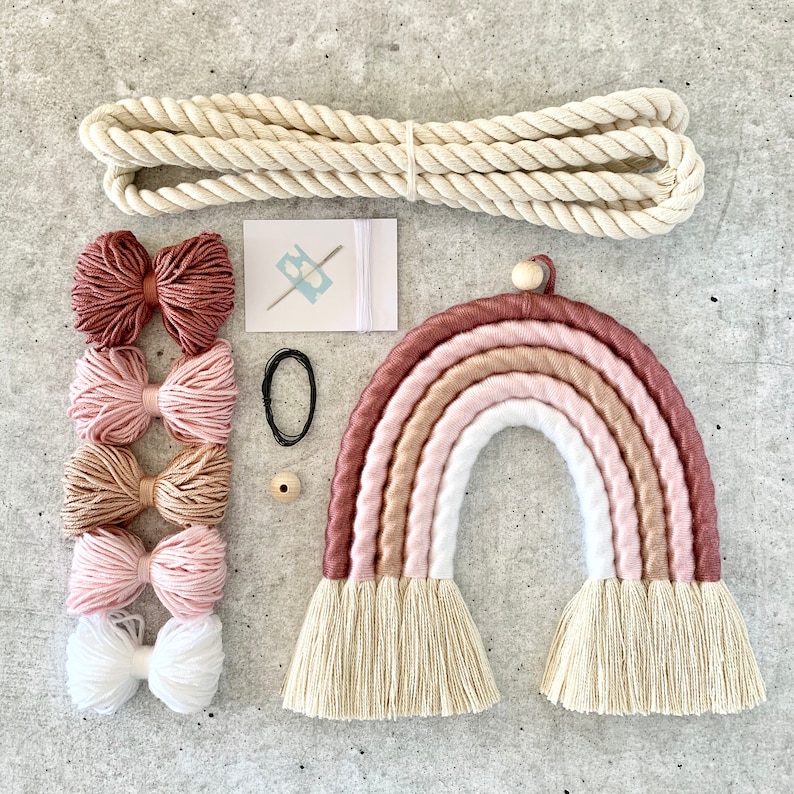 Op de afbeelding: Een DIY regenboog wanddecoratie set met een houten kraal, wit touw, roze, beige en bruin garen en een naald. De set bevat ook een witte kaart met de tekst "BE" en een zwart elastiekje.