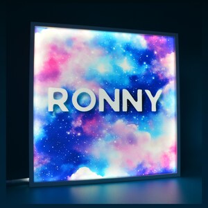 May include: A square, illuminated sign displaying the name "RONNY" in white block letters. The background features a colourful nebula with pink, blue, and white clouds and stars. The sign is framed in white.