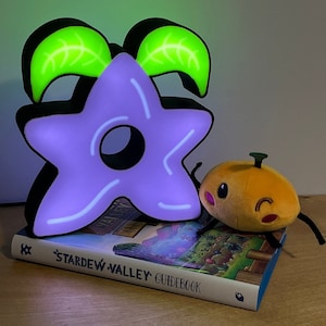 Könnte beinhalten: Ein lilafarbenes sternförmiges Nachtlicht mit grünen Blättern, eine orangefarbene Plüschfigur mit einem lächelnden Gesicht und ein Buch mit dem Titel "Stardew Valley Guidebook".
