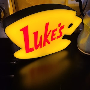 May include: A yellow and red illuminated sign shaped like a coffee cup with the text "Luke's" in red.