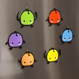 May include: Six colorful magnetic characters with a cartoon-like design. The characters are green, red, blue, purple, orange, and yellow. Each character has a small antenna on top of its head and two arms that are raised in the air. The characters are all smiling and have large eyes.