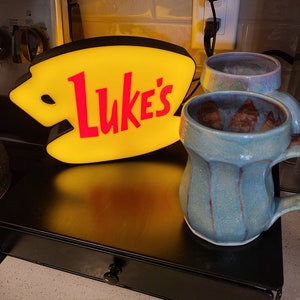May include: A yellow and red neon sign with the text "Luke's" on a black shelf with two ceramic mugs. One mug is blue and brown, the other is purple and white.
