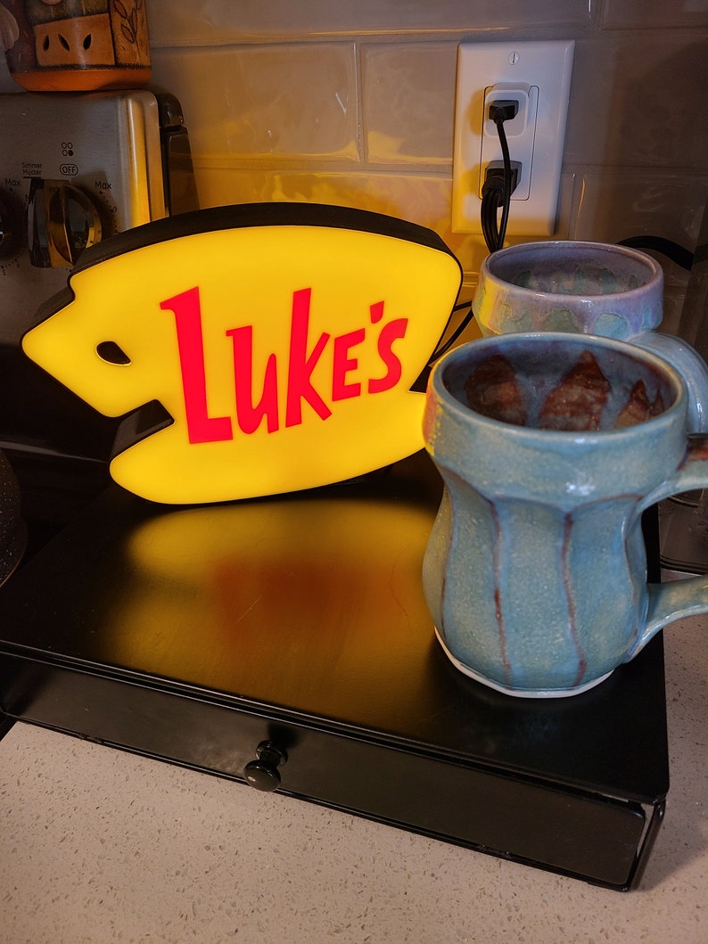 May include: A yellow and red neon sign with the text "Luke's" on a black shelf with two ceramic mugs. One mug is blue and brown, the other is purple and white.