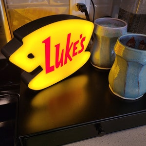 May include: A yellow and black diner sign with the word "Luke's" in red letters. The sign is lit up and is sitting on a black counter next to two ceramic mugs.