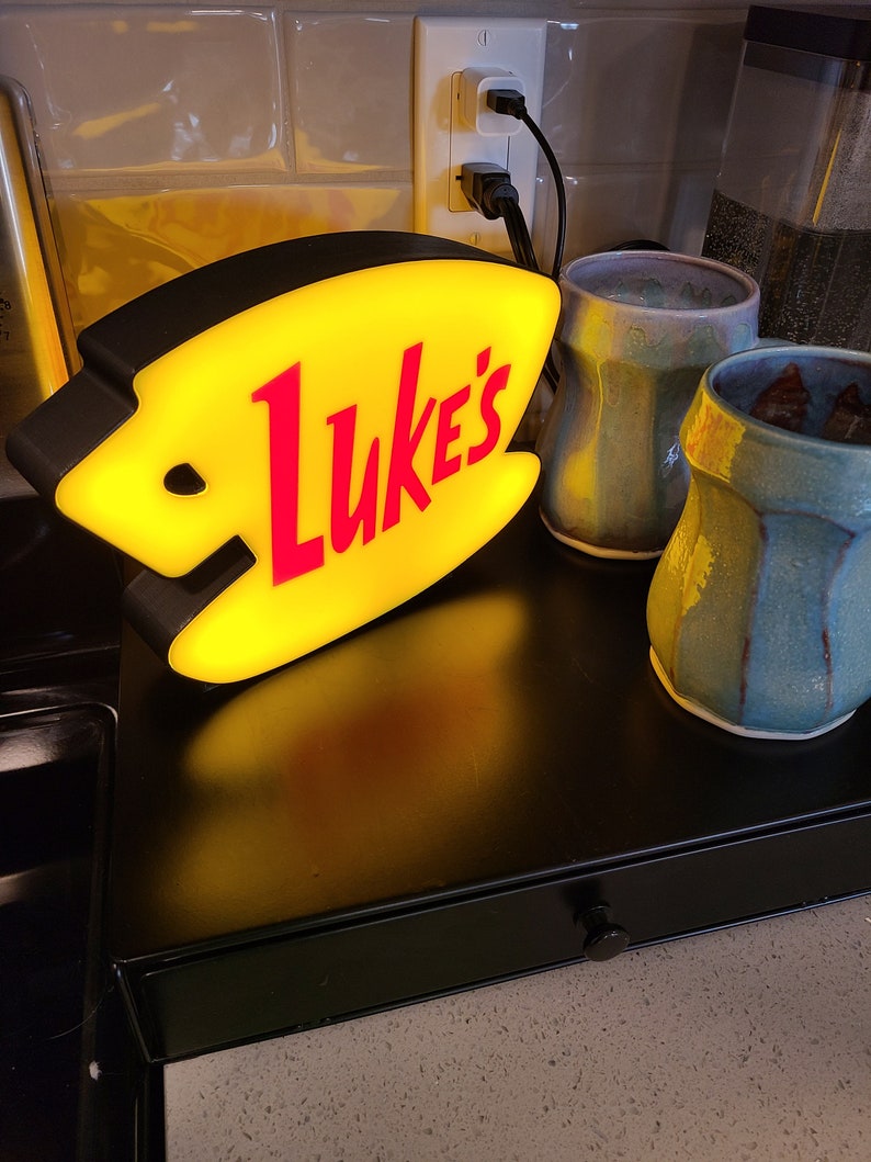 May include: A yellow and black diner sign with the word "Luke's" in red letters. The sign is lit up and is sitting on a black counter next to two ceramic mugs.