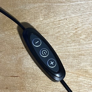 May include: Black remote control with three buttons: a minus sign, a power button with a circle and a plus sign. The remote is connected to a black cord.