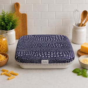May include: A square fabric bowl cover with a dark blue background and a white pattern of circles and crosses. The cover features an elasticized light gray hem and a small white rectangular tag labeled Practical Homestead on the front edge.