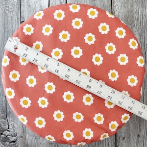 Könnte beinhalten: Ein rundes, korallenfarbenes Stoffstück mit einem sich wiederholenden Muster aus weißen Gänseblümchen mit gelben Zentren. Ein Lineal liegt quer über dem Stoff und misst etwa 33 cm. Geeignet für Näharbeiten oder Bastelprojekte.