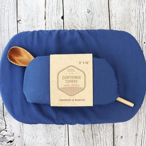 Puede incluir: Un juego de fundas azules para recipientes con una cuchara de madera. El juego incluye una funda enrollada y una funda plana. La etiqueta dice "Container Covers" y "Handmade in Winnipeg". La funda enrollada mide 23 cm x 33 cm.