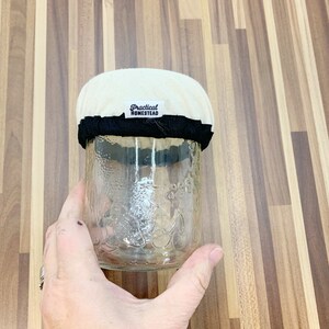 May include: A clear glass jar with a cream fabric top and a black band. The jar features a floral pattern and the text "Practical Homestead" on the fabric. The jar is held by a hand.