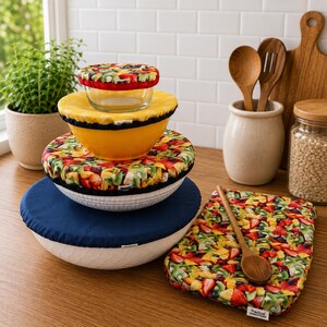 May include: A stack of four bowls covered with fabric bowl covers in various patterns including a fruit print and solid navy blue. A rectangular fabric cover with a fruit print lies flat next to the bowls, featuring a small white tag with text.