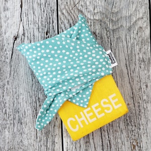 May include: A teal and white polka dot fabric cheese wrap covers a yellow wooden cheese block. The word "CHEESE" is printed in white on the block. The tag reads "Practical Homestead". The wrap is tied in a knot.