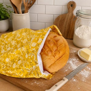 May include: A round loaf of golden-brown bread sits partially inside a yellow fabric bag featuring a white floral pattern. The bag has a plain white interior lining and is resting on a wooden cutting board next to a silver bread knife.