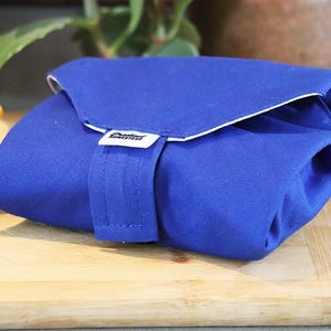 Può includere: Una borsa per il pranzo in tessuto blu con finiture bianche e chiusura con cinturino. La borsa è appoggiata su un tagliere di legno accanto a un panino.