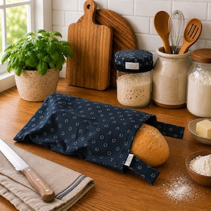 May include: A dark blue fabric bread bag with a repeating small circular pattern and a white tag that reads Practical Homestead, containing a loaf of crusty bread. The bag features a fold-over closure and is made of a durable, woven textile material.