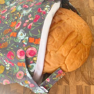 May include: A gray bread bag with a floral pattern, holding a loaf of bread. The bag's fabric features pink, orange, and yellow flowers. The bread is golden brown and partially visible.