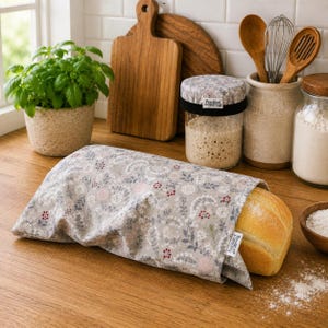 May include: A rectangular fabric bread bag featuring a light grey background with a delicate floral pattern in shades of pink, red, and blue. The bag is partially open, revealing a golden-brown loaf of bread inside with a small white label attached.