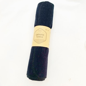 May include: A rolled set of six black unpaper towels, held together by a brown paper band. The band features the text "UNPAPER TOWELS" in a stylized font, with the words "SET / 6" in the upper right corner.