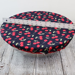 May include: A navy blue bowl cover with a red cherry pattern. The cover is approximately 15 inches in diameter. The edge of the cover is red. The cover is designed to fit over a bowl.