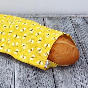 May include: A yellow fabric bread bag featuring a repeating pattern of small black and yellow bees with white wings. The bag is open at one end, revealing a golden-brown loaf of bread inside. A small white tag reads Practical Homestead.