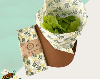 May include: Two white fabric food wraps with a green and yellow leaf pattern. The wraps are made by Slow & Co. and are labeled "Emballages alimentaires réutilisables - Food Wraps". One wrap is folded into a bowl shape and filled with green lettuce. The other wrap is flat and lying on a brown surface.