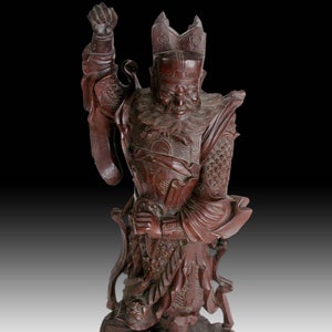May include: A dark brown wooden sculpture of a warrior figure standing on a base with intricate carvings. The warrior is wearing a crown and armor, and is holding a sword in his raised right hand.