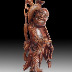 Peut inclure: Une statue en bois sculptée à la main représentant un Bouddha souriant, debout sur une base aux détails complexes. La statue est en bois foncé et a une couleur brun rougeâtre chaude.