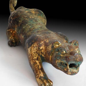 May include: Bronze sculpture of a crouching feline with a gold and verdigris finish. The cat is depicted in a stalking pose, with its mouth slightly open. The sculpture has a detailed texture and is set against a dark background.